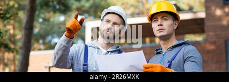 Building ideas. Portrait of two builders looking away while standing outdoors with an open blueprint discussing the half completed cottage house Stock Photo