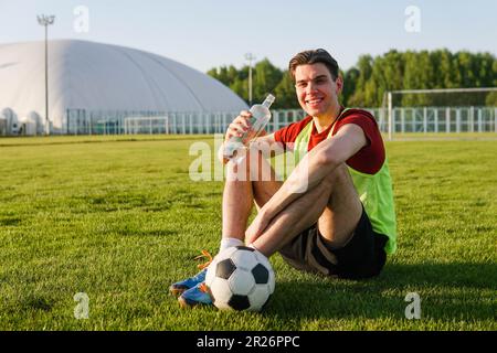 Young man football player resting after training sitting on grass and drinking water from a bottle. Soccer ball on the field Stock Photo