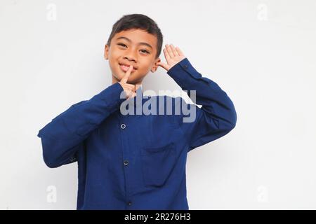 Smiling confident boy wearing a blue casual shirt looking at the camera while put fingers on mouth and put hand on the ear for sign just listening no Stock Photo