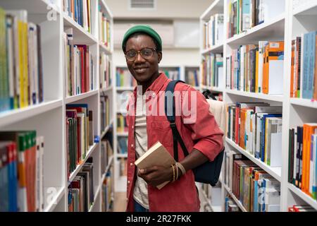 Smiling african american man student holds book in university library between bookshelves. Stock Photo
