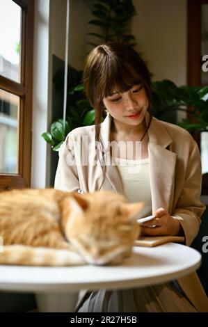 Attractive young Asian woman sits and reads a book at a table by the window while her chunky orange cat is sleeping on a table. Stock Photo
