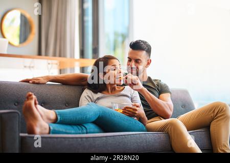Love, happy couple on sofa and eating chips in living room of their home. Caring or bonding together, relaxing or comfortable and happy married people Stock Photo