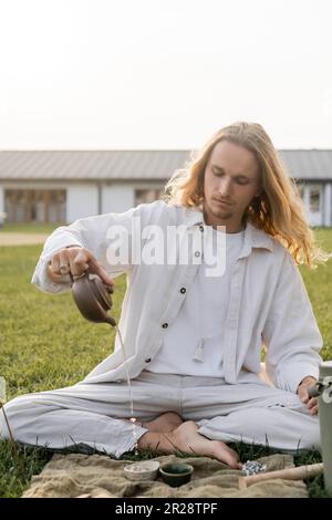 young man in white clothes pouring hot water from clay oriental teapot during traditional ceremony in countryside Stock Photo