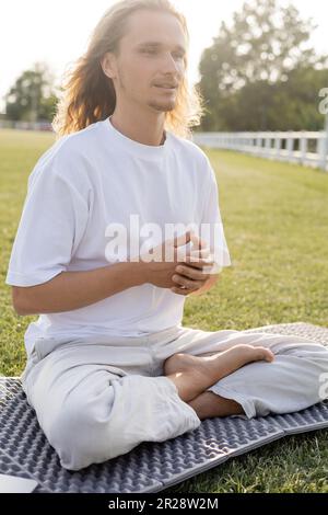 full length of barefoot man in white t-shirt and cotton pants sitting in easy pose and meditating on grassy field Stock Photo