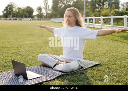 young man sitting in easy pose with outstretched hands during online yoga lesson on laptop outdoors Stock Photo