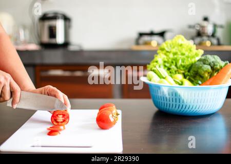 Close-up, A woman's hand was holding a knife and cutting tomatoes on a cutting board in the home kitchen. Cucumbers, lettuce, carrots in blue containe Stock Photo