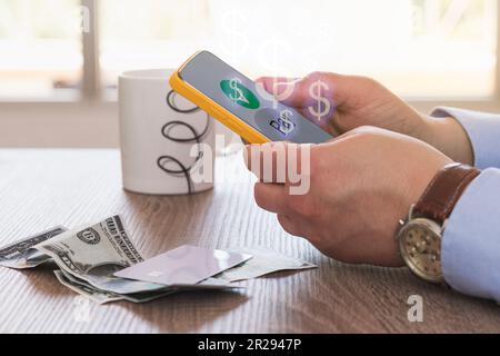 Crop anonymous male with wrist watch sitting at wooden table near money and white cup while using mobile app to purchase online Stock Photo