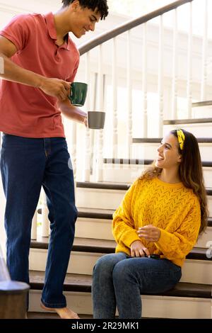Happy diverse couple having coffee, sitting on stairs at home Stock Photo
