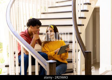 Happy diverse couple sitting on stairs at home talking and using tablet Stock Photo