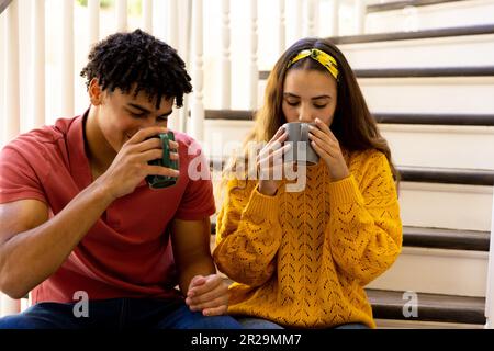 Happy diverse couple sitting on stairs at home drinking coffee Stock Photo