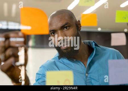 Bearded african american businessman sticking adhesive notes on glass wall in office Stock Photo
