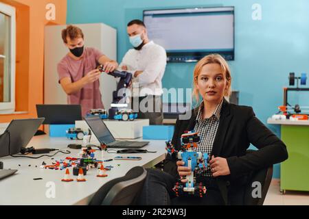 A woman sitting in a laboratory and solving problems and analyzing the robot's verification. In the background, colleagues are talking at an online Stock Photo