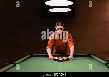 Sports game of billiards on a green cloth. Caucasian woman collect multi colored billiard balls in the form of a triangle with numbers shoot. Sports Stock Photo