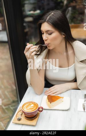 young woman with long hair eating cheesecake next to cup of cappuccino on bistro table while sitting in leather jacket next to window inside of modern Stock Photo