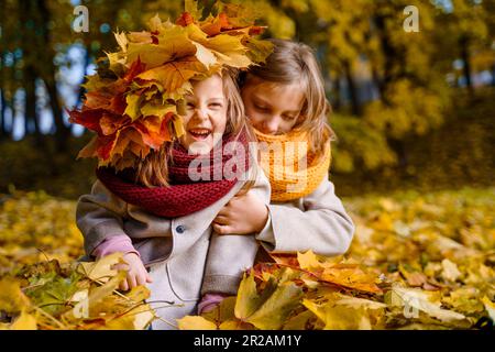 Beautiful little girls in bright yellow red orange autumn warm knitted scarf snood with head wreath of maple leaves,foliage walking in forest. Fun pla Stock Photo