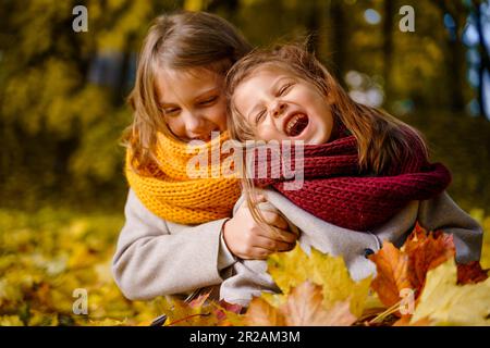 Beautiful little girls in bright yellow red orange autumn warm knitted scarf snood with head wreath of maple leaves,foliage walking in forest. Fun pla Stock Photo