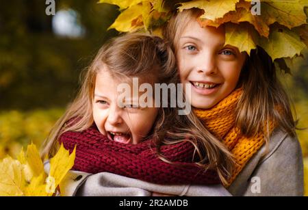 Beautiful little girls in bright yellow red orange autumn warm knitted scarf snood with head wreath of maple leaves,foliage walking in forest. Fun pla Stock Photo