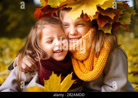 Beautiful little girls in bright yellow red orange autumn warm knitted scarf snood with head wreath of maple leaves,foliage walking in forest. Fun pla Stock Photo