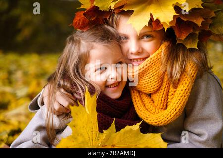 Beautiful little girls in bright yellow red orange autumn warm knitted scarf snood with head wreath of maple leaves,foliage walking in forest. Fun pla Stock Photo