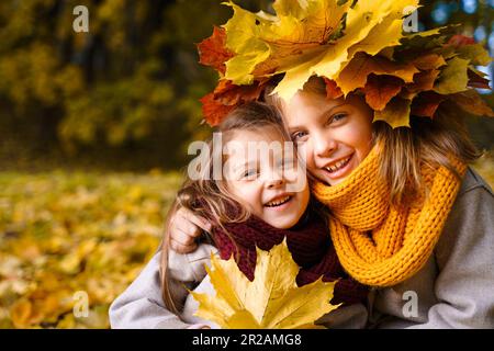 Beautiful little girls in bright yellow red orange autumn warm knitted scarf snood with head wreath of maple leaves,foliage walking in forest. Fun pla Stock Photo