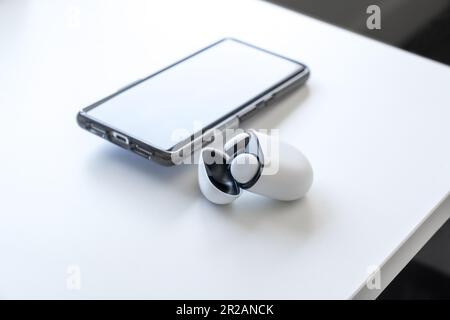 Wireless earbud headphones in their case, with the lid open, sitting on a white desk with a smartphone in the background. Room for copy space. Stock Photo
