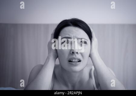 Black and white image. A frightened young woman is sitting on the sofa and holding her head with her hands. the concept of a panic attack. Stock Photo