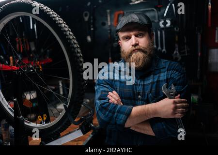 Portrait of bearded cycling mechanic male holding wrench in hand standing by bicycle in repair workshop with dark interior, looking at camera Stock Photo