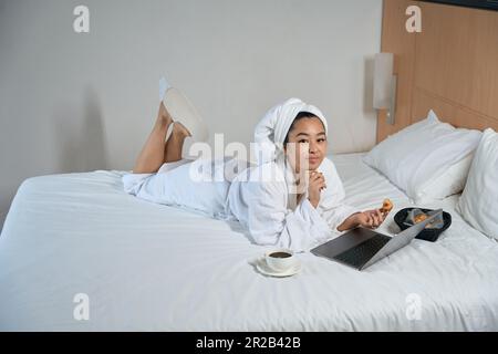 Woman in bathrobe using laptop is having breakfast on bed Stock Photo