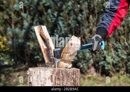 Close-up of split wood logs by sharp axe and human hand in working glove. Male arm holding splitting ax with plastic handle at firewood preparation. Stock Photo