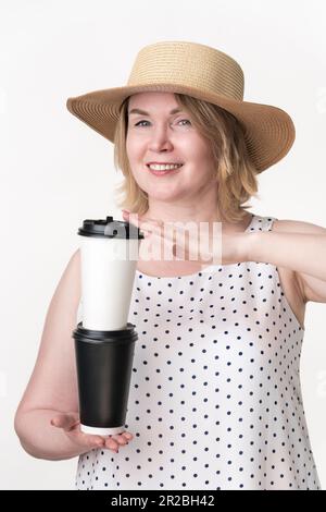 Happy smiling female holding both hands white and black disposable paper coffee cups stacked one on top of other. Woman 49 years old in summer dress Stock Photo