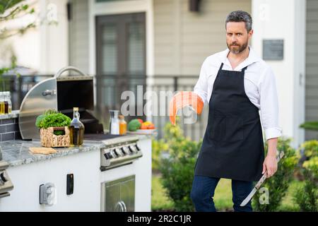 Handsome man in shirt and in cook apron preparing fish on barbecue. Grilling salmon outdoor. Male cook cooking salmon fillet on grill. Fish for barbec Stock Photo