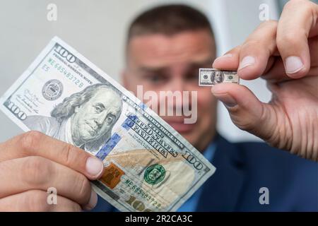 young businessman guy holds a small bill in his hand as a symbol of declining income. Small salary concept. A sad face . dollar inflation Stock Photo