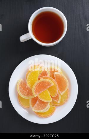Marmalade in shape of citrus fruits slices and cup of tea on black wooden background, top view Stock Photo