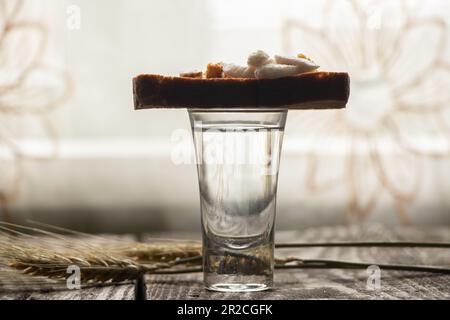 a glass of vodka next to a piece of black bread with bacon on a wooden table in the kitchen for lunch in Ukrainian Stock Photo