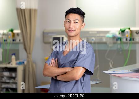 Portrait of happy asian male doctor in scrubs crossing arms and smiling at hospital Stock Photo