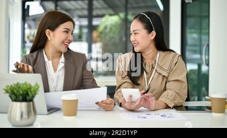 Two cheerful and attractive millennial Asian businesswomen or accountants are working and analyzing financial data reports in the office together. Stock Photo