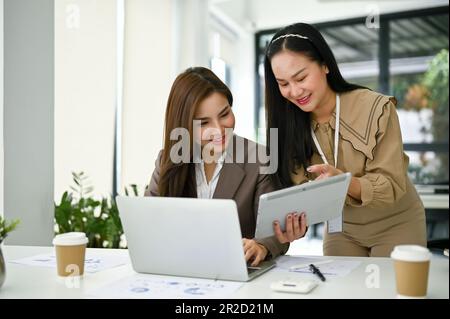 A professional millennial Asian female office worker showing something on tablet screen to her colleague, discussing and planning a new project in the Stock Photo