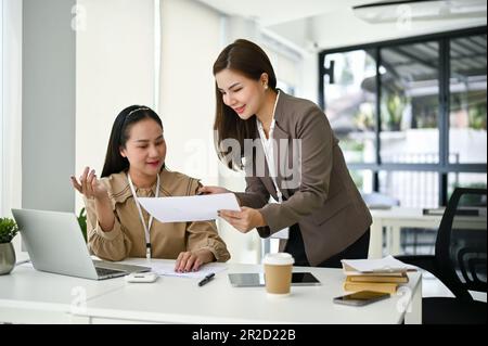 A beautiful young Asian female secretary is showing something on document to her boss, listening to her boss brief her on a new project, and working i Stock Photo