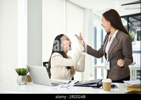 Two cheerful and smiling millennial Asian businesswomen are giving high fives to cheer each other up or celebrating their project success. Stock Photo