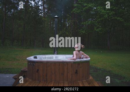 Young woman chilling in hot tub surrounded by relaxing forest and green nature Stock Photo