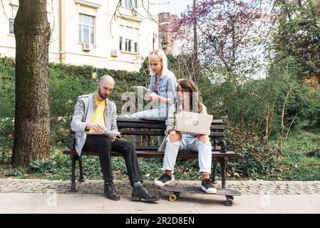 Diverse hipster happy students studying outdoors with different electronic devices, sitting together on bench at university campus. Freelancers busine Stock Photo