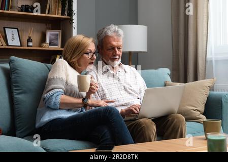 Happy middle aged family couple relaxing on sofa, using computer web surfing information shopping together at home. Elderly man showing laptop apps to Stock Photo