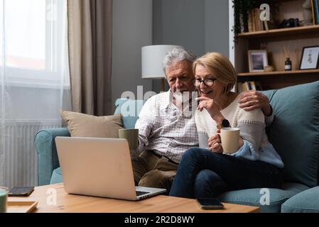 Smiling older wife and husband hugging, using laptop, sitting on cozy couch, watching movie or shopping online, looking at screen, mature man and woma Stock Photo