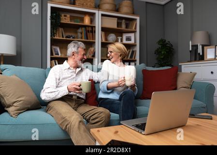 Smiling older wife and husband hugging, using laptop, sitting on cozy couch, watching movie or shopping online, looking at screen, mature man and woma Stock Photo