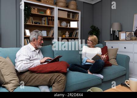 Happy senior married couple relaxing together at home with the wife sitting back from her husband as she reads a book while he surfs the internet on a Stock Photo