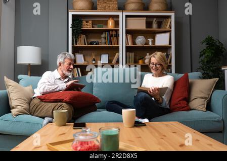 Simple living. Happy middle-aged senior couple 70s husband and wife sit relax on couch in living room reading book drinking tea together, calm elderly Stock Photo