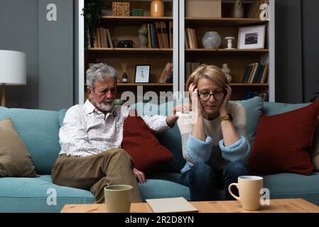 Sad mature woman sitting away from her husband after having quarrel. Senior couple not talking having argue at their home. Marriage crisis relationshi Stock Photo