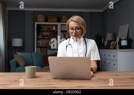 Serious female doctor using laptop and writing notes in medical journal sitting at desk. Senior woman professional medic physician wearing white coat Stock Photo