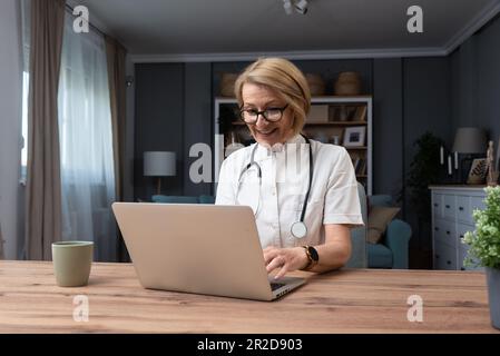 Serious female doctor using laptop and writing notes in medical journal sitting at desk. Senior woman professional medic physician wearing white coat Stock Photo