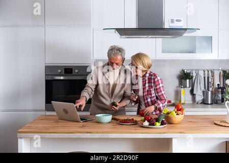 Happy old middle aged senior couple using laptop computer preparing healthy food diet vegetable salad at home together, searching recipes, ordering sh Stock Photo